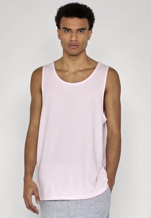 Homme aux cheveux bouclés portant un débardeur rose clair et un pantalon gris, debout devant un fond gris uni.