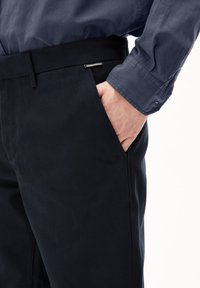 Zwarte broek gemaakt van een zachte, gestructureerde stof. Kenmerkt zich door een brede tailleband, zijzak en een ARMEDANGELS-label op de riem.