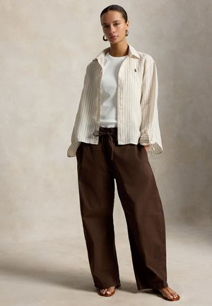 Γυναίκα που στέκεται, φοράει λευκό τ-shirt, μπεζ ριγέ ανοιχτό πουκάμισο, φαρδιά σκούρα καφέ παντελόνια και καφέ σανδάλια, μπροστά σε ουδέτερο φόντο.