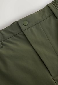 Olivgrüne Hose mit flacher Vorderseite, vertikaler Rippstruktur und grünem Knopfverschluss am Bund.