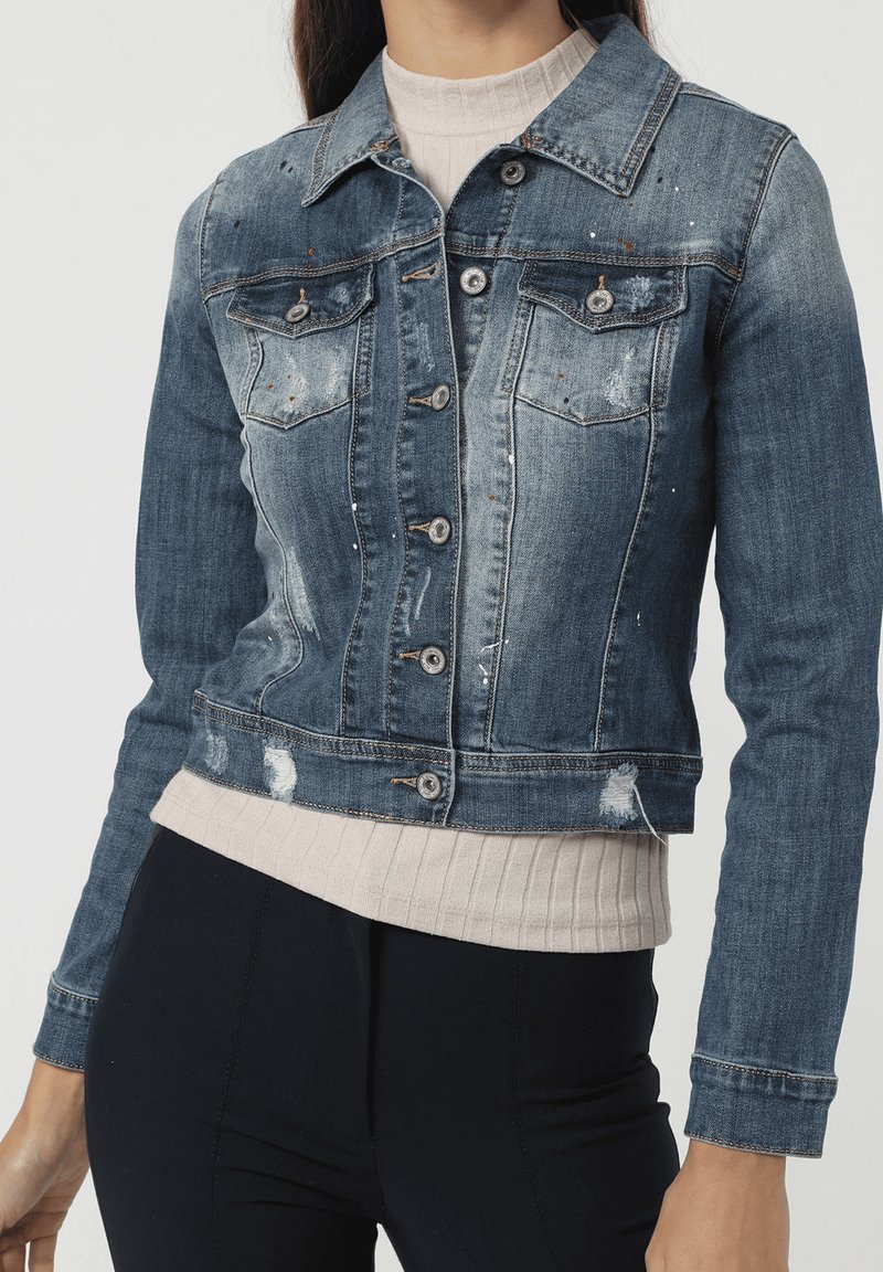 Chaqueta de denim en azul medio con detalles desgastados, cierre de botones, dos bolsillos en el pecho y un largo recortado, superpuesta a una blusa de punto beige.