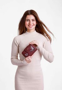 Hnědá kožená peněženka s designem motýlů, texturovaným povrchem a klopovým zavíráním. Model ji drží proti jednobarevnému pozadí.