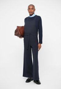 Navy jumpsuit with adjustable straps, wide legs, and a belt detail. Paired with a light blue shirt and a brown leather handbag.