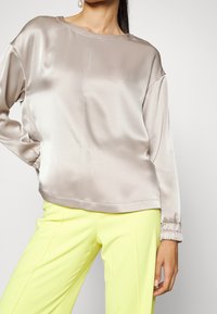 Femme portant un top à manches longues en satin gris clair avec des poignets élastiques et un pantalon jaune vif sur un fond uni.