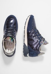 Un paio di sneakers blu navy con tessuto lucido e glitterato, suole bianche e accenti sulla linguetta iridescenti, su uno sfondo bianco.