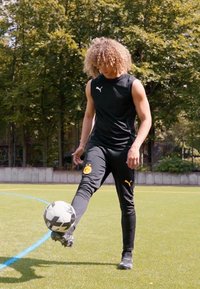 Zwarte mouwloze sporttop, zwarte broek met geel logo. Speler vaardigheden met een zwart-witte voetbal op een groen veld.