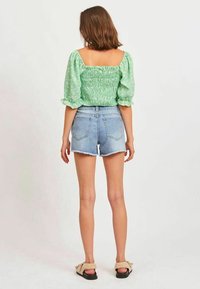 Zielona, kwiecista bluzka z marszczeniami i bufkami, zestawiona z jasnoniebieskimi szortami dżinsowymi z postrzępionymi brzegami oraz brązowymi klapkami.