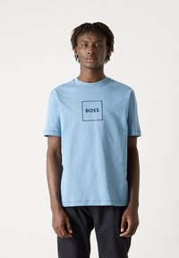Camiseta de algodón de color azul claro con un logo cuadrado y el texto "BOSS" en azul oscuro. Mangas cortas y un cuello redondo clásico.
