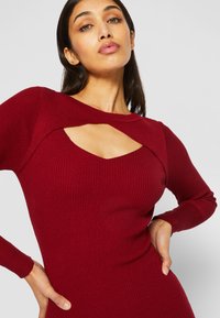Abito a costine borgogna con design a taglio su scollatura e spalle, maniche lunghe e silhouette aderente.