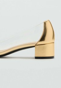 Chaussure plate en métal doré avec un talon carré, des côtés transparents et une texture lisse. Caractérisée par un bout rond et des accents de semelle noire.