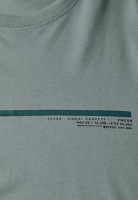 T-shirt en coton vert clair avec un col rond et une rayure horizontale bleu canard. Le texte imprimé inclut des coordonnées géographiques et une marque.