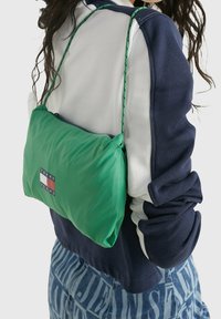 Zelená nylonová taška přes rameno se zipovým uzávěrem a nastavitelným popruhem, s logem v červené, bílé a modré barvě, vystavená na tmavě modrém topu.
