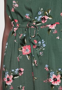 Grön blommig klänning med rosa, röda och blå blommor. Har en rynkad midja med ett knytband, tillverkad av ett mjukt och slätt tyg.