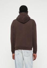 Barna kapucnis pulóver nagy kapucnival, esőfedett vállakkal és bordázott mandzsettával. Világoskék farmernadrággal párosítva, hátsó zsebekkel.