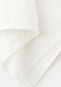 Tissu blanc doux avec des textures délicates tricotées et tissées, superposées et pliées sur un fond blanc.