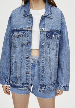 Femme portant une veste en denim à lavage acide surdimensionnée avec des détails de boutons, associée à un short en denim taille haute et un crop top blanc.