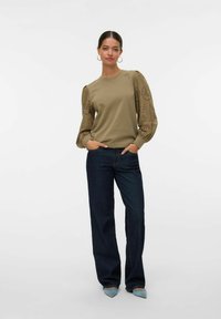 Sudadera de un verde oliva claro con un intrincado diseño de recortes en las mangas, combinada con unos vaqueros de pierna ancha de color azul oscuro y unos zapatos de punta azul claro.