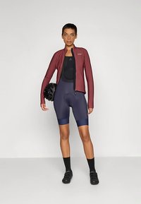 Žena stojaca v bordovom cyklistickom bundu, čiernom cyklistickom tričku, tmavomodrých cyklistických šortkách, čiernych ponožkách, topánkach, držiaca čiernu prilbu.