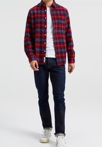 Camicia di flanella a quadretti rossi e blu con maniche lunghe, chiusura con bottoni frontale e taschino sul petto, abbinata a jeans di denim scuri e sneaker bianche.