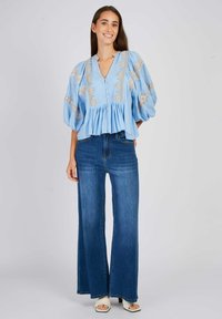 Lichtblauwe blouse met opstaande mouwen en geborduurde details, gecombineerd met high-waisted blauwe flared jeans en witte open-toe sandalen.