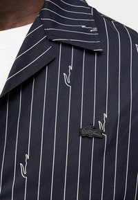 Πουκάμισο ναυτικού μπλε με λευκές ρίγες, διακοσμημένο με κεντημένα λογότυπα κροκόδειλου και γυριστό γιακά. Λείο ύφασμα με δομημένο σχέδιο.