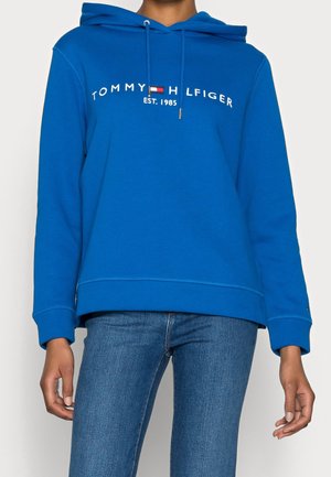 Blauwe hoodie met tekst "Tommy Hilfiger EST. 1985" en logo over de borst, gedragen met een blauwe spijkerbroek door een persoon die tegen een effen achtergrond staat.