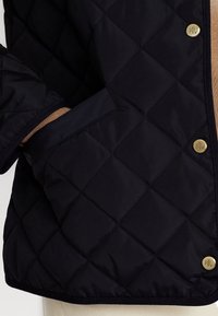 Tmavě modrá prošívaná bunda s kosočtvercovým vzorem, patentkami se zlatými akcenty a boční kapsou, vyrobená z hladkého, voděodolného materiálu.