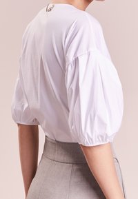 Blusa bianca con maniche a sbuffo, collo rotondo e una spilla decorativa sul retro. Il tessuto appare liscio e leggero. Indossata con una gonna grigia.