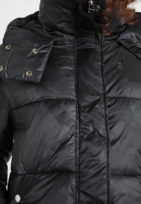 Černá péřová bunda s vysokým límcem, s knoflíky na ramenech a matnou texturou. Má viditelné logo a prošívaný vzor.