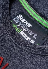 Šedá texturovaná tkanina s černou nálepkou s nápisem "Super Drysport", doplněná zeleným a šedým prošíváním. Obsahuje červené písmo dole.