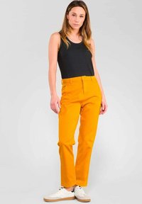 Gelbe Baumwollhosen mit gerader Passform, Knopfverschluss und Gürtelschlaufen, kombiniert mit einem schwarzen Tanktop und weißen Turnschuhen.