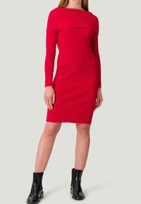 Robe rouge côtelée avec un décolleté asymétrique et des manches longues, présentant une silhouette ajustée, complétée par des bottines noires à la cheville.