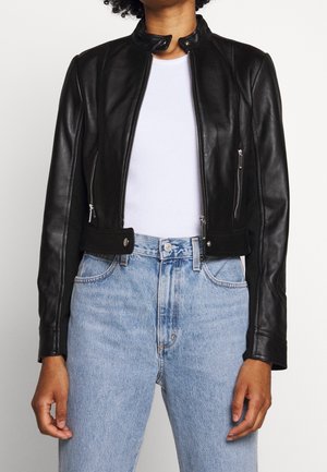 Persoon die een aansluitend zwart leren jack draagt over een wit overhemd en lichtblauwe high-waisted jeans tegen een effen achtergrond.