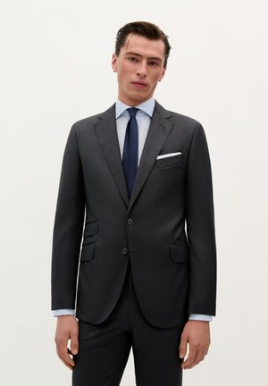 Traje gris oscuro con solapas de pico, diseño de un solo botón, cierre de dos botones y bolsillos laterales. Camisa de vestir azul claro y corbata azul marino.