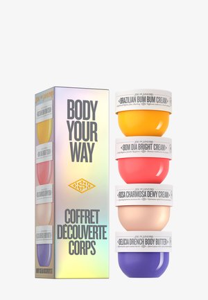 Quattro barattoli di crema impilati, colorati e etichettati con i nomi dei prodotti, accanto a una scatola con scritto "Body Your Way" e testo in francese su sfondo bianco.