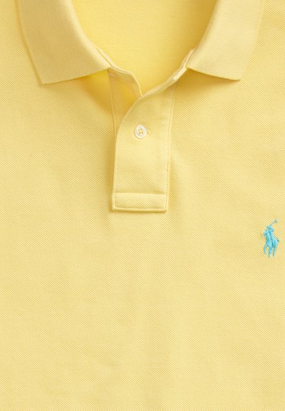 Geltonas polo marškinėliai su apykakle, vienu užsegtu mygtuku ir mažyliu mėlynu siuvinėtu arkliuko logotipu dešinėje pusėje.