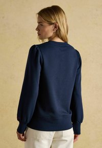 Tumši zils krekls ar garām puffed piedurknēm, apaļu kakla izgriezumu, ribotām aprocēm un jostu. Sarkana teksta detaļa pie aproces. Mīksta auduma tekstūra.