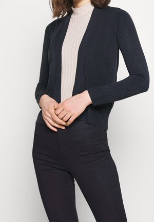 Kvinde iført en cremefarvet ribstrikket mock-neck top med en sort åbning-forrest cardigan og sorte tætsiddende bukser, hænderne foldet foran.