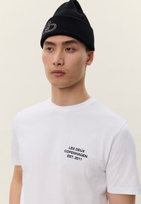 Jauns vīrietis, valkājot melnu cepuri un baltu T-kreklu ar uzrakstu "LES DEUX COPENHAGEN EST. 2011" uz krūtīm, uz vienmērīga fona.