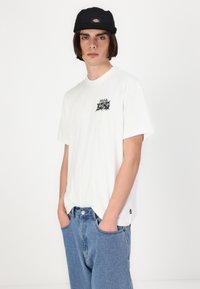 Hvid bomulds t-shirt med et sort grafisk design på venstre bryst. Sammen med blå denimbukser og en sort kasket. Enkel design.