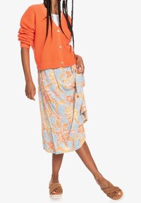 Orangefarbener Strickcardigan mit V-Ausschnitt und Holzknöpfen, kombiniert mit einem hellblauen, floral gemusterten Rock. Das Outfit wird durch braune Slipper-Sandalen abgerundet.