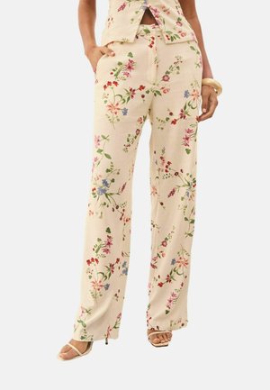 Pantaloni bej largi cu imprimeu floral colorat, purtați cu top asortat și pantofi cu toc decupați la degete, mâinile în buzunare și brățară la încheietură.