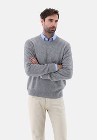 Grau gestrickter Pullover mit rundem Halsausschnitt, gerippten Bündchen und Saum, getragen über einem hellblauen Hemd, kombiniert mit beigen Hosen.