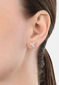 Pendientes de studs en tono dorado que presentan una piedra rectangular clara junto a una pequeña perla blanca, situados sobre el fondo de una oreja de tono piel.