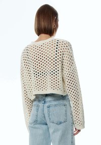 Häkelpullover in Off-White mit einem lockeren, cropped Design, das ein großes Lochmuster und weite Ärmel aufweist, kombiniert mit hellblauen Jeans.