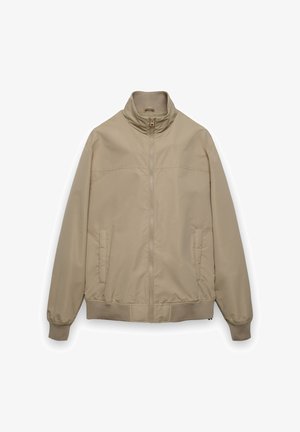 Beżowa kurtka na zamek z kołnierzem typu stójka, z bocznymi kieszeniami i prążkowanym obszyciem. Gładki, lekki materiał o subtelnym połysku.