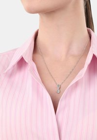 Srebrny naszyjnik o skręconym wzorze nieskończoności z małym skupiskiem przezroczystych cyrkonii. Noszony na różowej koszuli w paski z kołnierzykiem.