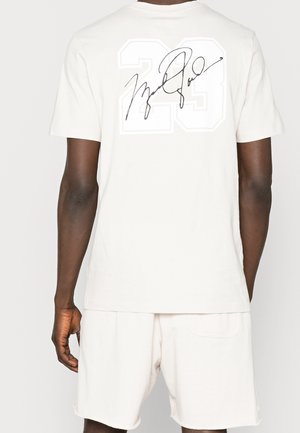 Person iført en hvid t-shirt og shorts, set bagfra, med stort nummer 23 og sort kursiv signatur på t-shirtens bagside.