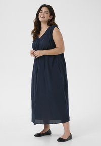 Marineblaues ärmelloses Maxikleid mit gerafftem Ausschnitt, seitlichen Taschen und fließender Silhouette. Getragen mit schwarzen Ballerinas. Schlicht und lockerer Schnitt.