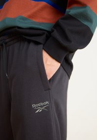 Crne trenirke s džepovima i elastičnim pojasom, s Reebok logotipom i kontrastnim gornjim dijelom u smešnim, zelenim i plavim prugama.